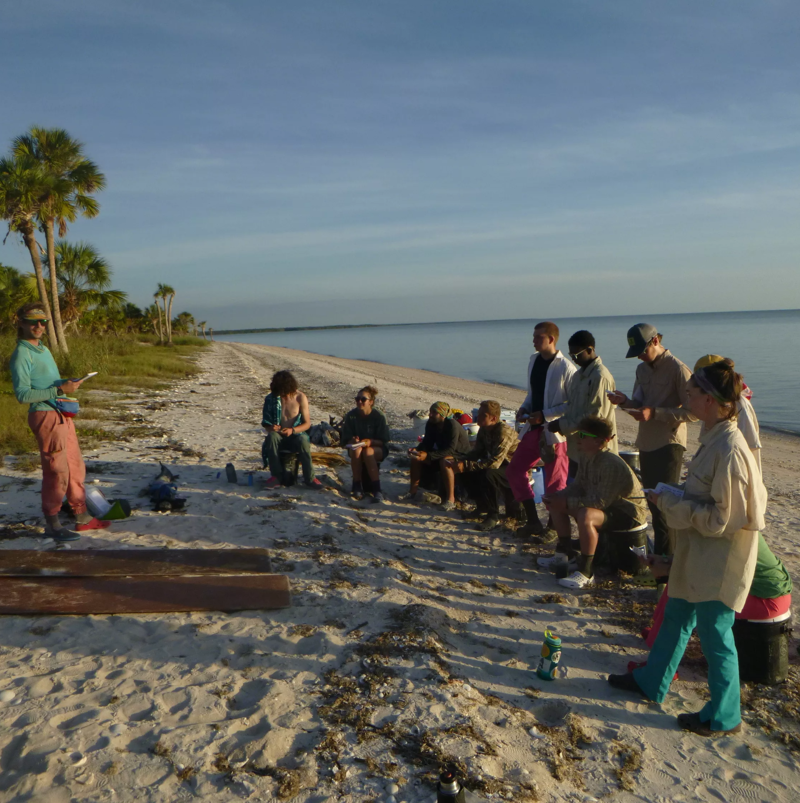 A group of people are gathered on a sandy beach, possibly for a lecture or discussion. They are sitting or standing, facing a person who appears to be speaking or presenting from a notebook. The beach is lined with vegetation, and the ocean is visible in the background under a clear sky. The scene suggests an outdoor educational or recreational activity.